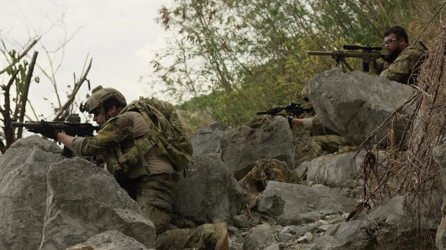 A team of special forces soldiers takes cover behind rugged rocks, conducting a tactical mission with a sniper providing overwatch in a hostile environment.