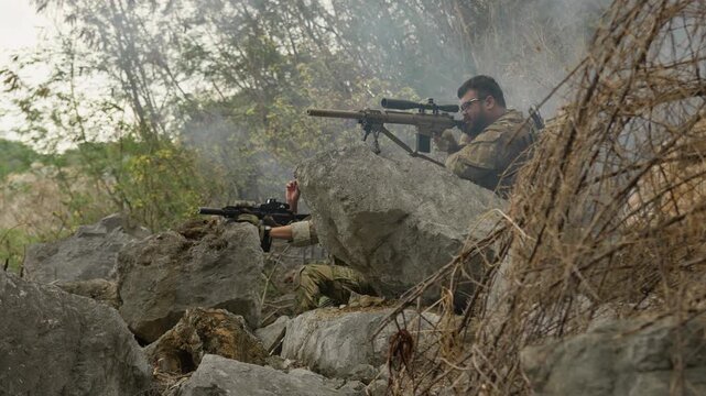 A military sniper team in camouflage takes up a defensive position among rocks, conducting surveillance during a tactical special forces operation in the jungle.