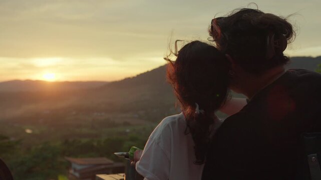 A mother and daughter watch the sunset over mountains from a viewpoint, sharing a quiet moment of family bonding and appreciating the tranquil scenic view.