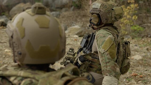 Two soldiers in full combat gear strategize during a mission, using tactical communication equipment to coordinate their next move in a rugged outdoor terrain.