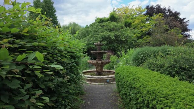 POV walking through lush garden path toward stone fountain. Peaceful park landscape, greenery and garden design concept.