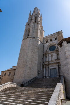 The towering spire and intricate masonry of the Basilica de Sant Feliu dominate the skyline in Girona's old quarter, viewed from the base of an ancient, wide stone stairway.