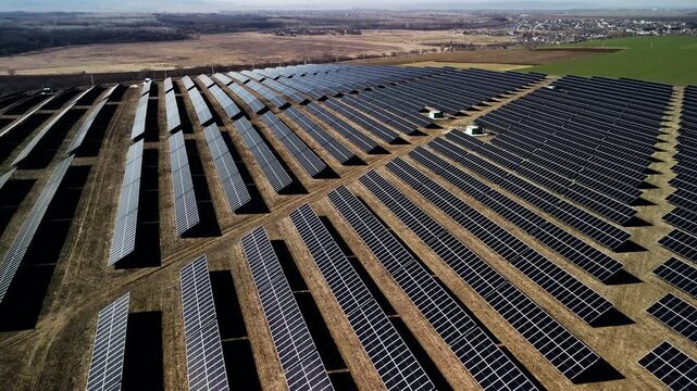 Aerial view of expansive solar panel farm with rows of photovoltaic panels arranged in a grid pattern under clear blue sky and green fields in the background