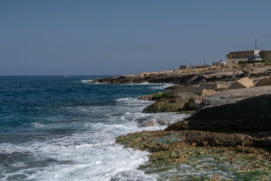 Rugged limestone coastline in Malta with turquoise Mediterranean waves breaking over algae covered rocks, capturing dramatic surf, coastal erosion, and natural seascape under a clear blue sky.