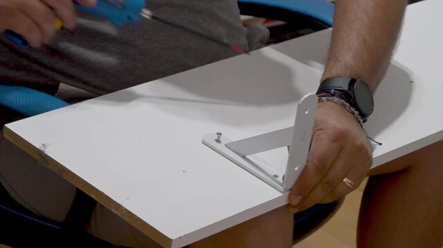 Close-up of a worker's hands attaching a metal bracket to a white wooden shelf with a power tool