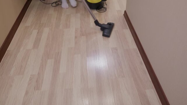 Person using yellow vacuum cleaner on light wood laminate floor during household cleaning routine.
