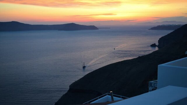 Colorful sunset sky above Santorini caldera with layered cliffs, calm Aegean Sea, and distant boats near Fira, Greece.