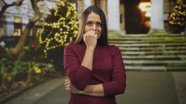 Woman biting fingernail with hand to mouth standing on building steps beside lit holiday tree and stone columns at evening; anxiety.