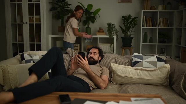 Lazy husband relaxing on the couch using his smartphone while his wife is cleaning the house, representing relationship problems