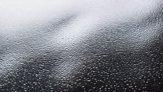 Slow pan closeup of water droplets sliding and coalescing on textured glass window surface in soft studio light macro reflective wet beads moving across windshield