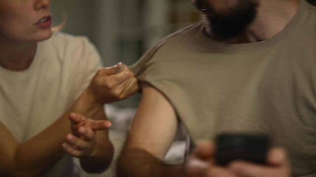 Angry woman grabbing her partner's shirt while he ignores her using his smartphone at home, relationship problems concept