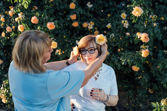 Playful Senior Woman Decorating Friend With Roses In Blooming Summer Garden