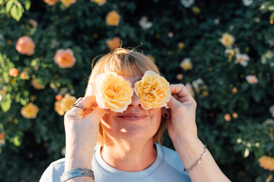 Cheerful Senior Woman Covering Eyes With Roses In Front Of Blooming Garden Wall