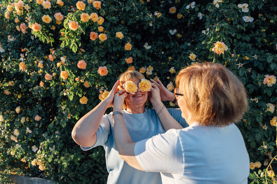 Playful Senior Woman Holding Yellow Roses Over Eyes In Blooming Summer Garden Portrait