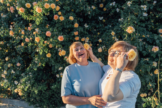 Cheerful Senior Friends Playing With Roses Near Blooming Garden During Summer Evening Walk