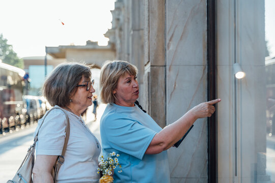 Helpful Senior Female Friends Looking At Store Window During Urban Shopping Walk