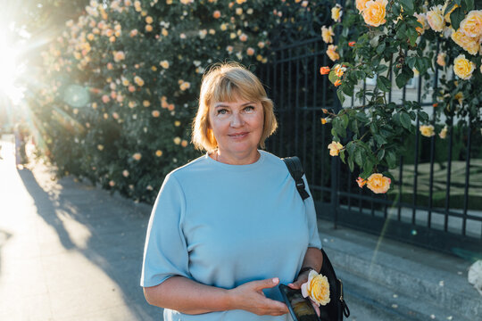 Beautiful Senior Woman Holding Smartphone Near Flowering Rose Wall In Evening Sunlight