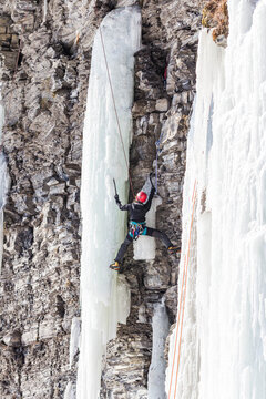 Ice climber ascending a frozen waterfall on a rocky cliff using ice axes and crampons