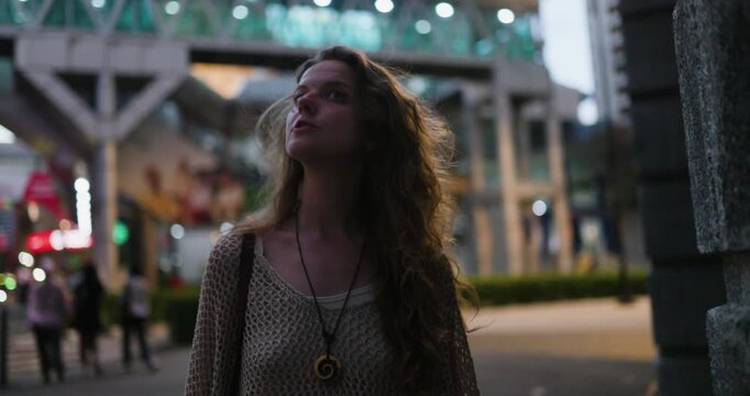 A thoughtful young woman with beautiful curly hair looks up at the urban skyline and city lights during an evening walk, reflecting on the modern world around her.