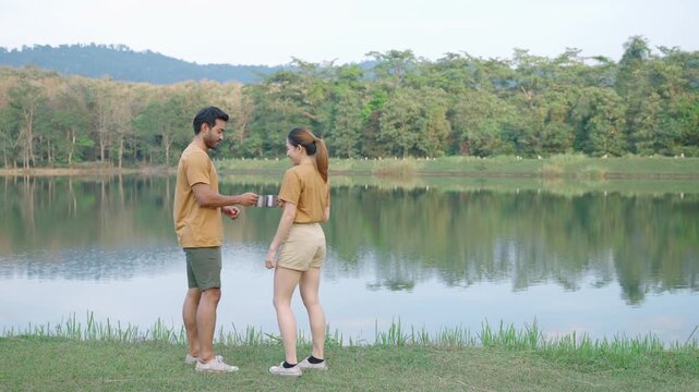 Man and woman holding metal mugs and sharing a coffee break by a calm lake surrounded by nature. Outdoor lifestyle, travel, friendship, companionship, and relaxation concept.
