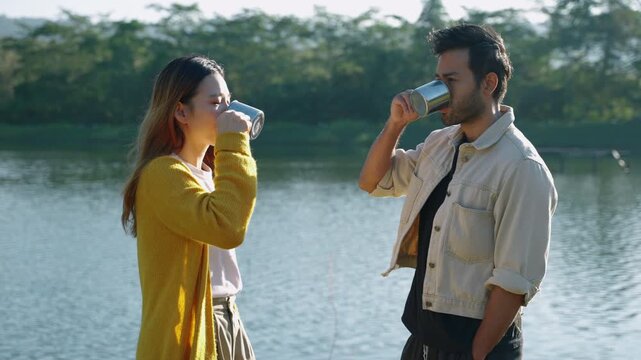 Man and woman holding metal mugs and sharing a coffee break by a calm lake surrounded by nature. Outdoor lifestyle, travel, friendship, companionship, and relaxation concept.

