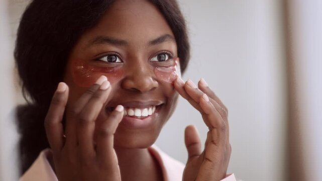 Smiling Young Black Woman Applying Under Eye Patches To Her Face In Front Of Mirror At Home, Engaging In Skincare Routine. Enjoying Self-care Moment In Relaxed Setting. Beauty Concept.