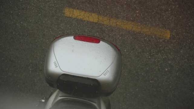 Silver hard scooter top case on road in rain. Rainstorm drops fall on plastic surface of parked motorcycle from top view.