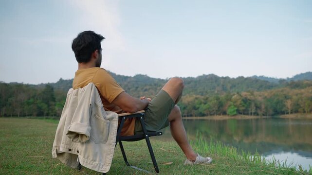 Young man sitting comfortably in a camping chair overlooking a peaceful lake and forest landscape. Outdoor recreation, mindfulness, freedom, relaxation, and nature retreat concept.
