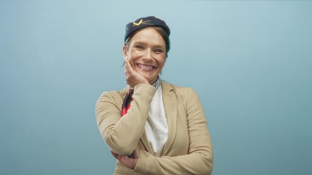 Woman flight attendant in uniform hat and neck scarf touching cheek while smiling in studio; approachable cheerful warmth.