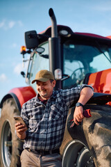 Naklejka premium Happy agriculturist using cell phone while leaning on tractor.