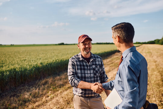 Happy mature farmer shaking hands with investment consultant in field.