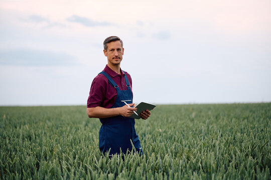 Agronomist using touchpad while inspecting wheat corps on his farm and looking at camera.
