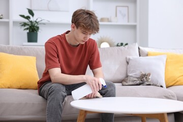 Fototapeta premium Housework. Teenage boy wiping white table with rag and bottle of detergent on sofa at home