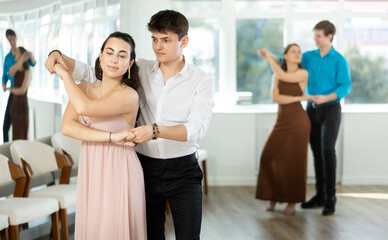 Fototapeta premium Expressive Hispanic guy in formal attire practicing sensual bachata with attractive young brunette in light flowing dress in dance class. Social dancing concept..