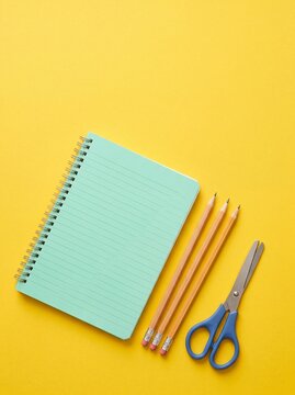 Mint Green Spiral Notebook with Three Pencils and Blue Scissors on Yellow Background