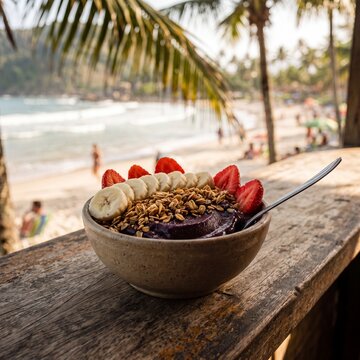 Fresh acai berry bowl topped with granola, banana slices, and strawberries on a rustic wooden ledge overlooking a sunny tropical beach