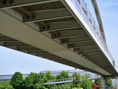 Low-angle, perspective 
view from underneath a sleek, modern 
pedestrian steel bridge. The 
architectural details show the metallic 
framework, beams, and safety mesh of 
the walkway. In the backgroun