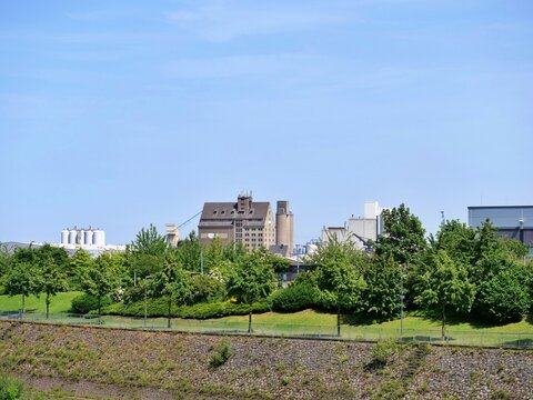  Wide shot of a historic 
brick grain silo and industrial factory complex rising behind a beautifully 
landscaped green park. The foreground 
features a stone-paved canal 
embankment with a safety fen