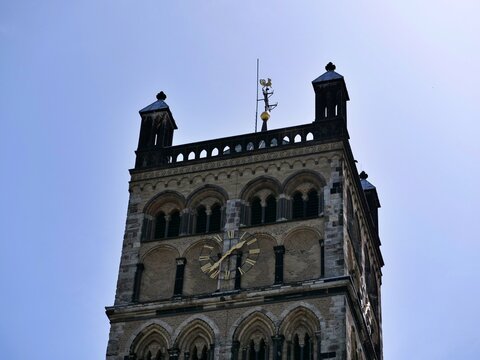 A detailed architectural 
photograph of the upper section of a 
historic Romanesque church tower. The 
ancient stone structure features 
traditional arched windows, decorative 
brickwork, and a promin