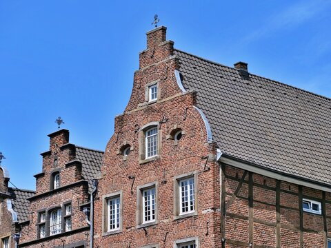 An architectural shot of a 
historic German brick building with a 
classic stepped or curved gable roof. 
The red brick facade features white
framed windows, decorative stone 
anchor irons, and a hist