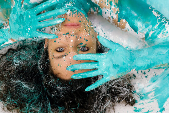 Close portrait of a woman covered in turquoise paint with splattered textures and expressive hands framing her face creating a vibrant artistic concept full of creativity and identity