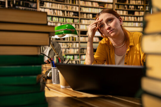 Stressed young woman rubbing forehead behind laptop at library table, framed by book stacks during focused exam preparation work, academic fatigue due to burnout, for study concepts.