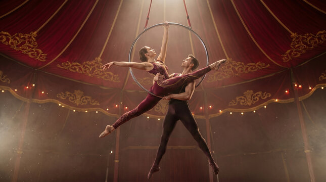 Professional aerial hoop artists performing dangerous acrobatic stunt under circus tent dome, man and woman in elegant matching costumes executing graceful routine on lyra ring 