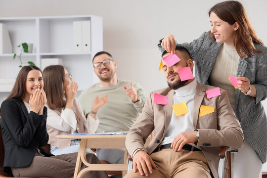 Office prank with sleeping colleague covered in sticky notes, humorous workplace interaction and team bonding, other team members in the background are laughing and enjoying the playful interaction