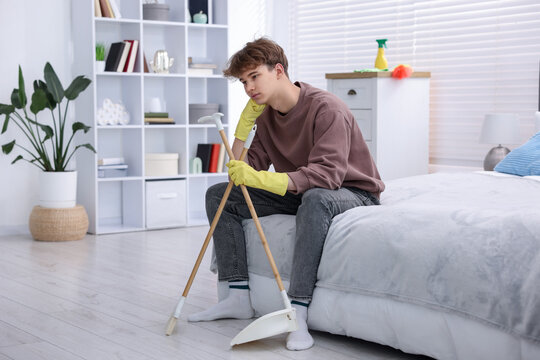 Housework. Tired teenage boy in rubber gloves with broom and dustpan sitting on bed at home