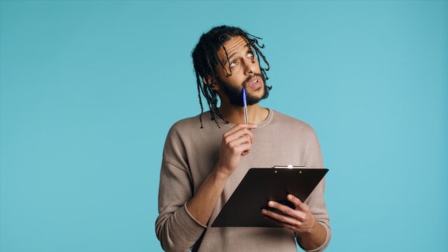 Creative man brainstorming ideas, writing on document using pen. BIPOC photographer using clipboard to complete shoot list for upcoming photography work, isolated over studio background, camera A