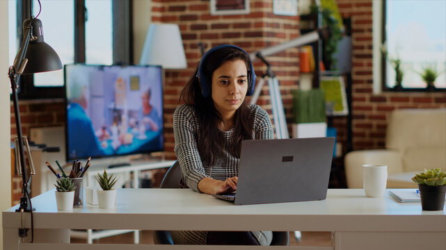 Relaxed cheerful remote worker listening to music on headphones while working from home. Smiling freelancer enjoying herself, typing on laptop in stylish personal office, camera B.