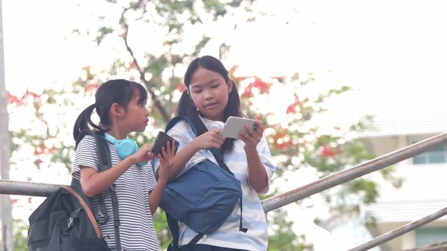 Cute young Asian school girls listening music via wireless headphones and digital tablet on campus step. Happy primary classmates enjoying e-learning back to school study loop  scene.