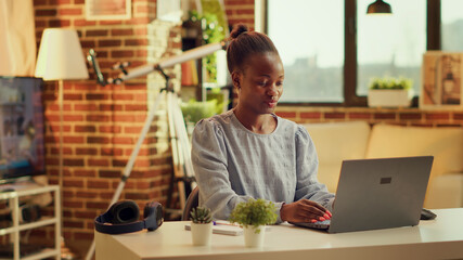 Fototapeta premium African American girl operates from home in the glow of sunset, publishing personal website article. Teleworker seeking to build online freelance business while refining abilities. Tripod shot.