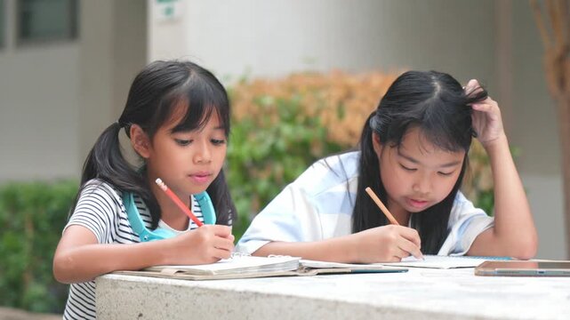 Cute young Asian school girls listening music via wireless headphones and digital tablet on campus step. Happy primary classmates enjoying e-learning back to school study loop  zone.